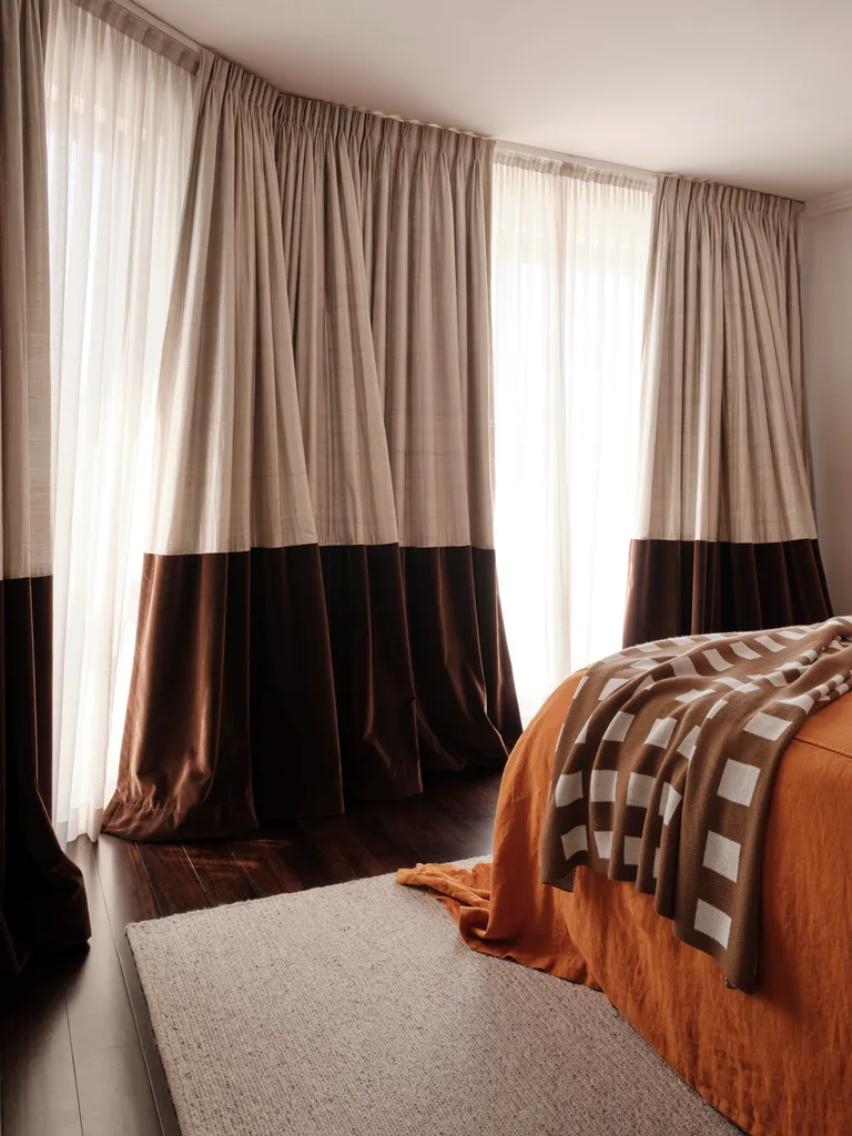 Bedroom with tall curtains, orange bedding, checkered blanket, wooden floor, and a light gray rug. It's the main bedroom in a family home.