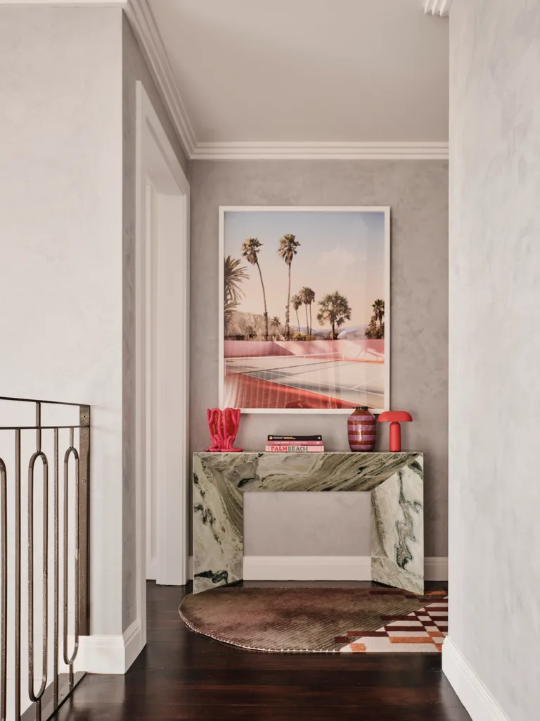 Decorative hallway with marble console table, red vase, lamp, and a framed palm tree photo above, wooden floor and neutral walls.