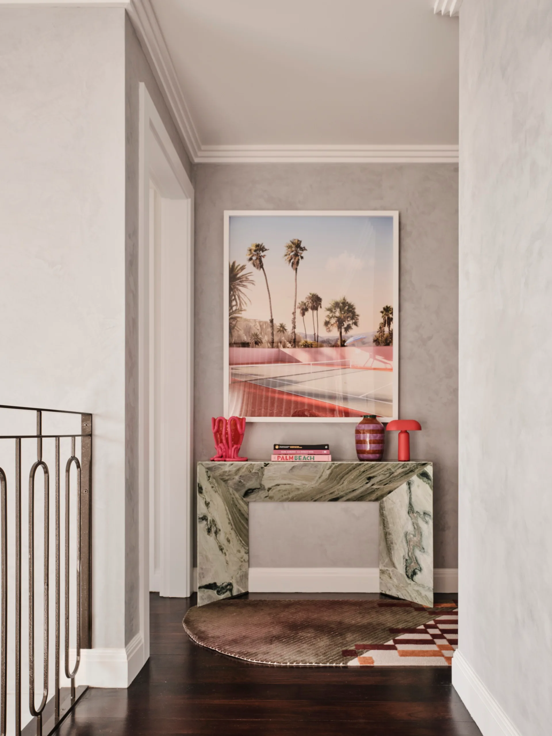 Decorative hallway with marble console table, red vase, lamp, and a framed palm tree photo above, wooden floor and neutral walls.