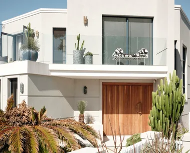 Modern house facade with large windows, wooden door, and lush greenery, including cacti and tropical plants.
