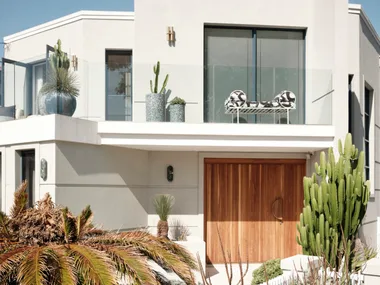 Modern house facade with large windows, wooden door, and lush greenery, including cacti and tropical plants.
