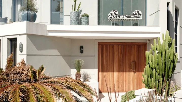 Modern house facade with large windows, wooden door, and lush greenery, including cacti and tropical plants.