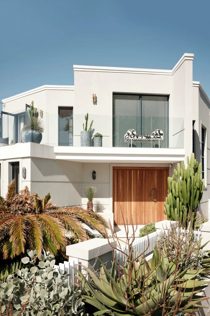 Modern house facade with large windows, wooden door, and lush greenery, including cacti and tropical plants.