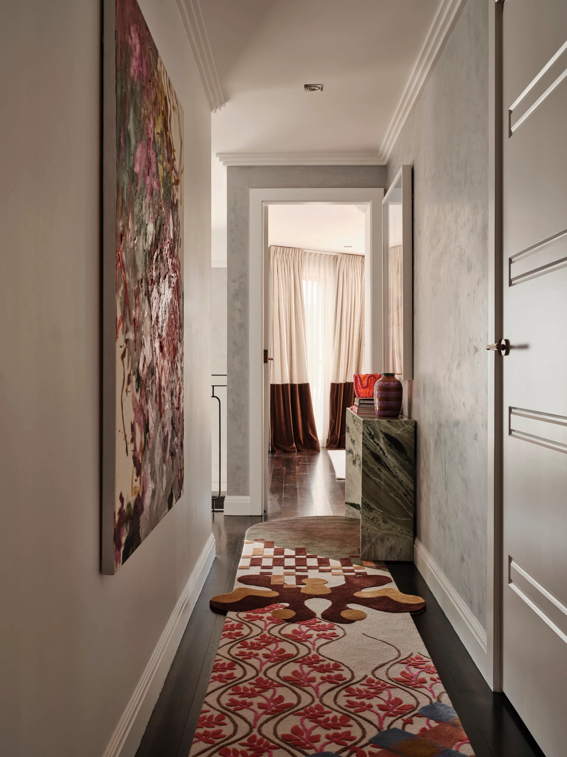 Narrow hallway with abstract artwork on the left wall, patterned carpet, and light-filled curtains at the end.