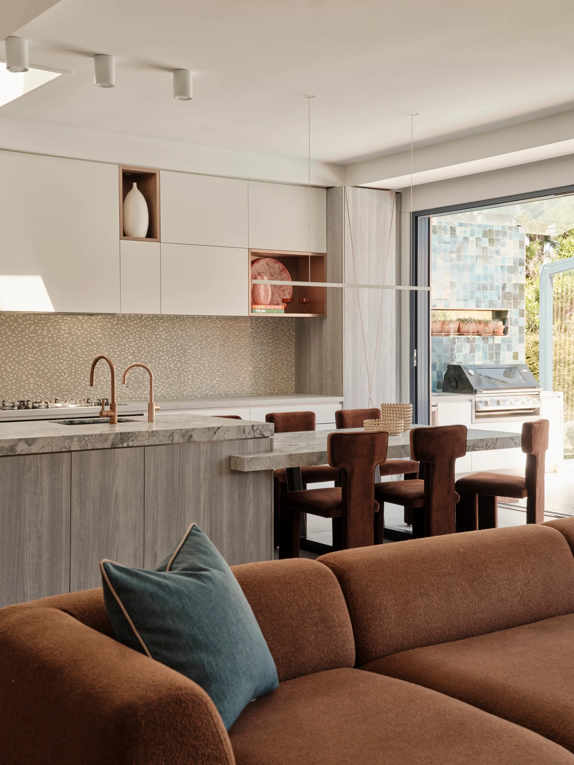 Modern kitchen with wood accents, marble counter, brown chairs, and a brown sofa with a blue cushion in the foreground.