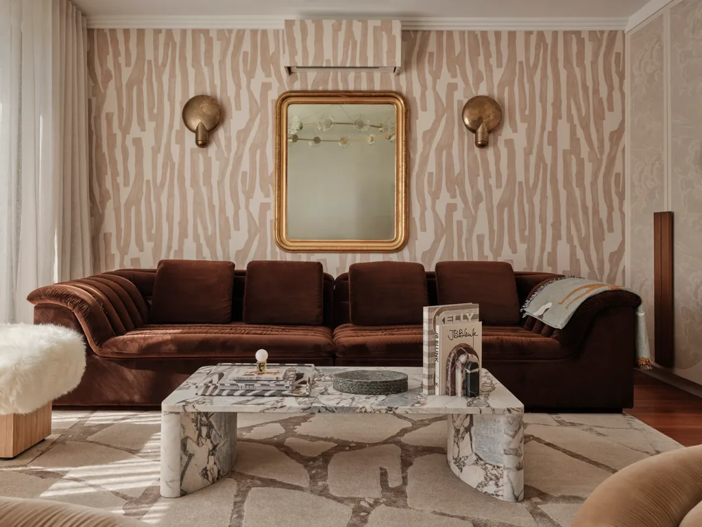 Brown couch in a living room with patterned wallpaper, marble coffee table, books, and decorative mirrors.