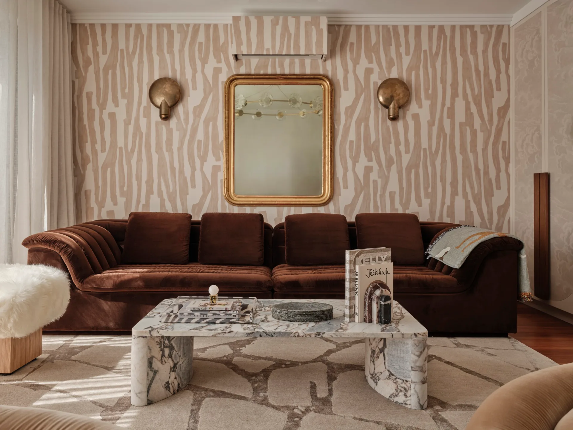 Brown couch in a living room with patterned wallpaper, marble coffee table, books, and decorative mirrors.