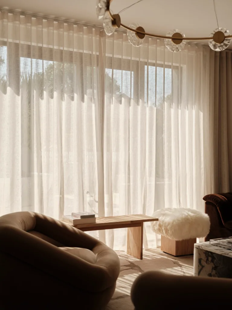 Cozy room with soft lighting through sheer curtains, featuring a plush chair, wooden table with books, and fluffy stool.