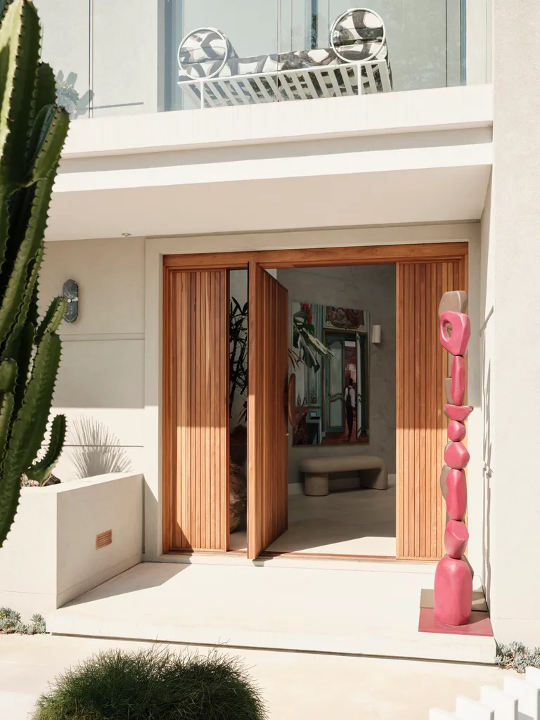 Entrance of a modern family home with wooden doors, red abstract sculpture, and a balcony above with a lounge chair.