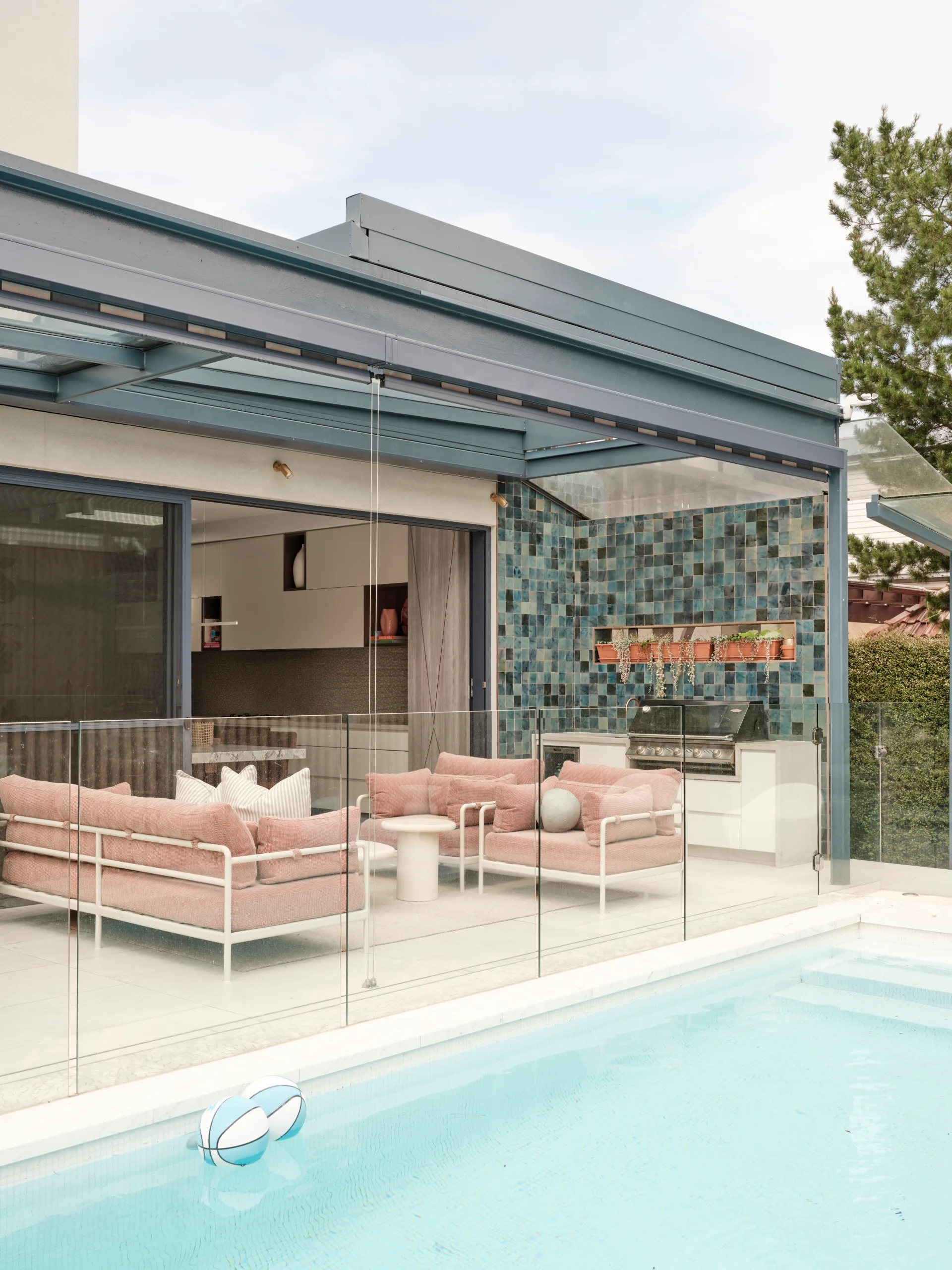 Outdoor kitchen and alfresco lounge with pink seating, coffee table, and a barbecue next to a swimming pool. Glass fencing surrounds the backyard area in this family home.