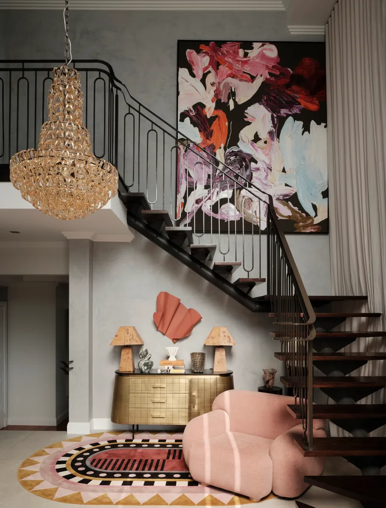 Luxurious interior with a chandelier, abstract art, a staircase, pink armchair, gold console table with drawers, and sculptural lamps. The abstract art is positioned above the staircase and is large in this family home.