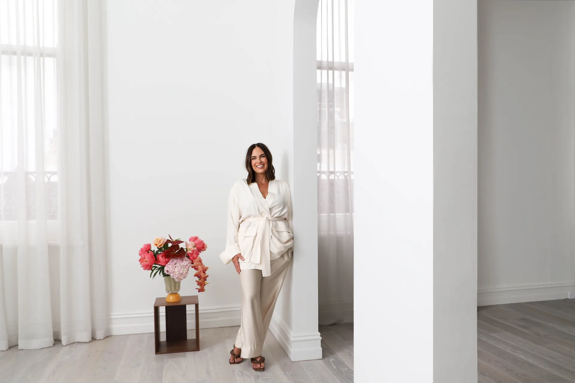 an Australian designer in white outfit smiling, standing by a white wall with flowers on a wooden table nearby.