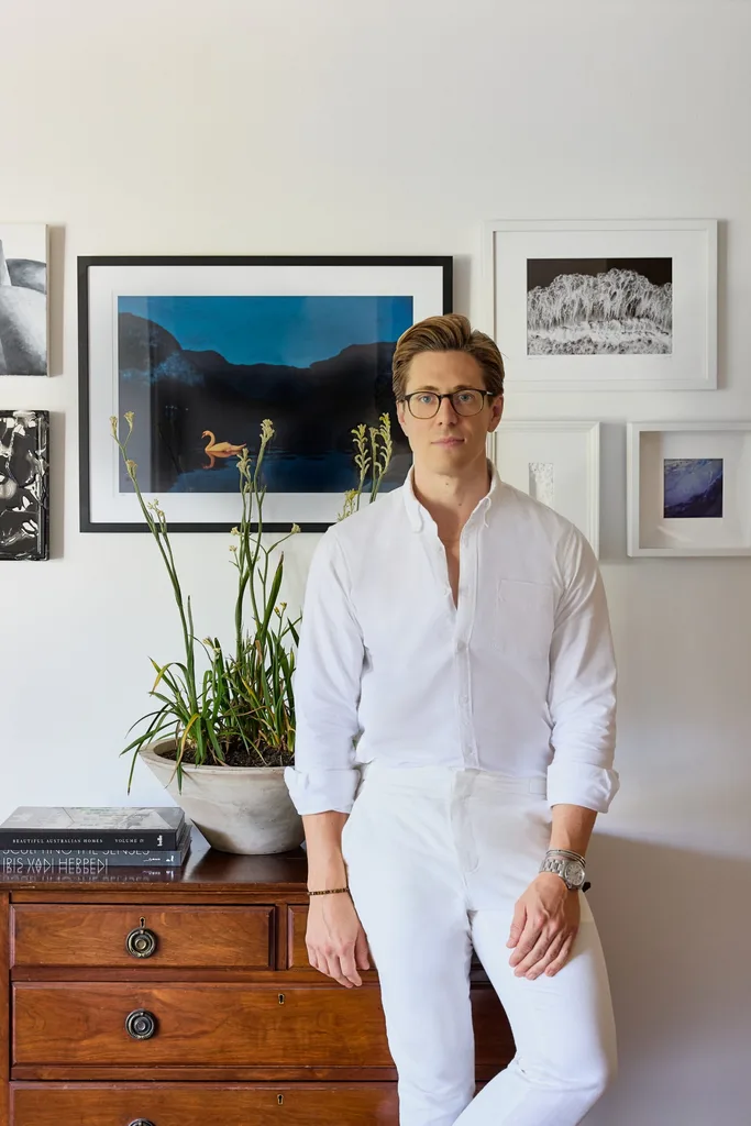 an Australian designer in white outfit leaning on a dresser with framed art and plant, against a white wall background.