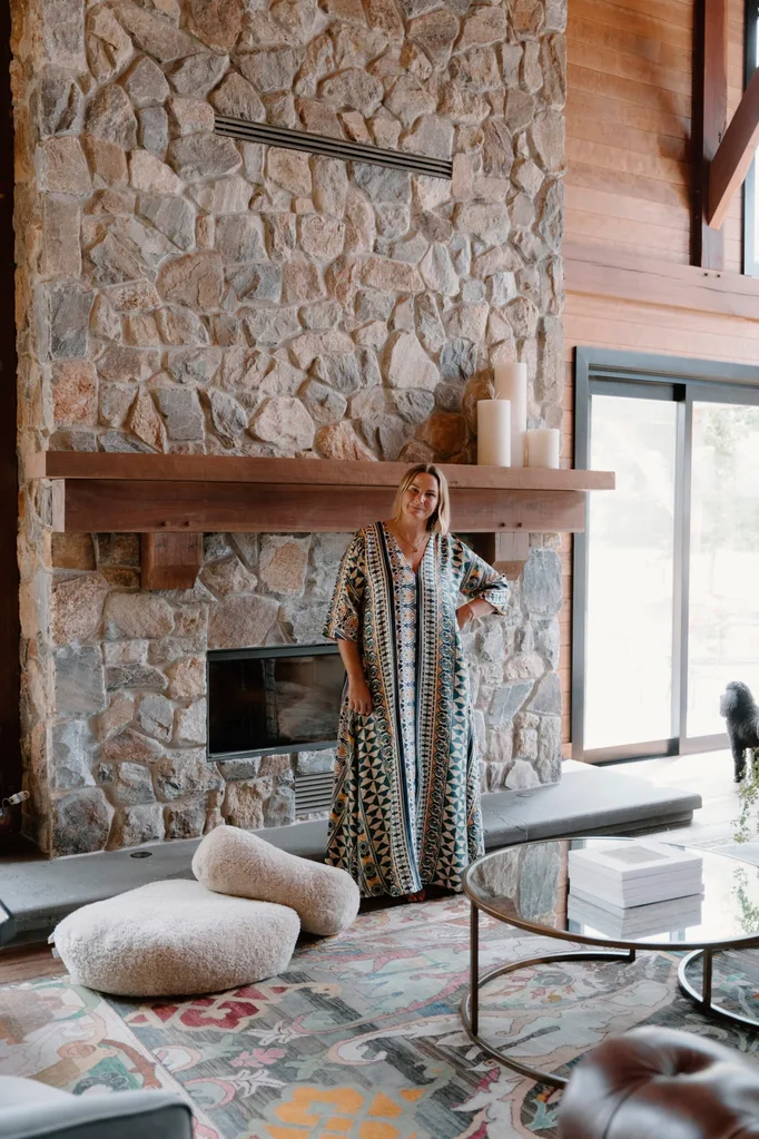 Woman, an Australian designer, in patterned dress standing by stone fireplace, with candles and glass table in a cozy living room setting.