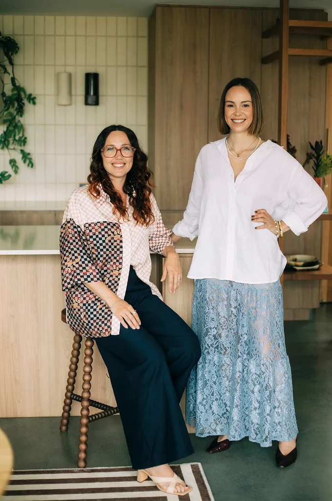 Two Australian designers smiling in a modern kitchen, one seated in a decorative top, the other standing in a white blouse and blue lace skirt.