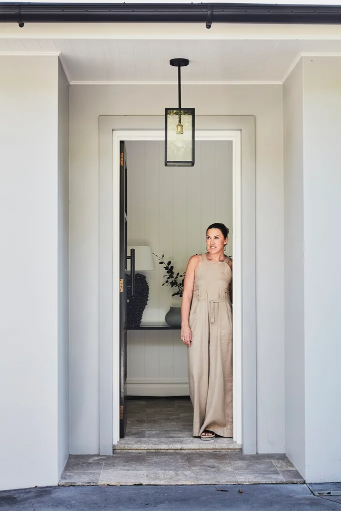 an Australian designer in a beige jumpsuit stands in a doorway, looking out with a light fixture above.
