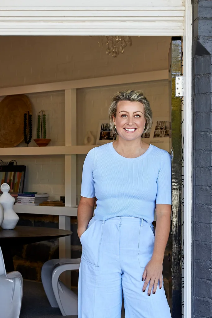 an Australian designer in a light blue outfit stands smiling at a doorway with a modern interior behind her.