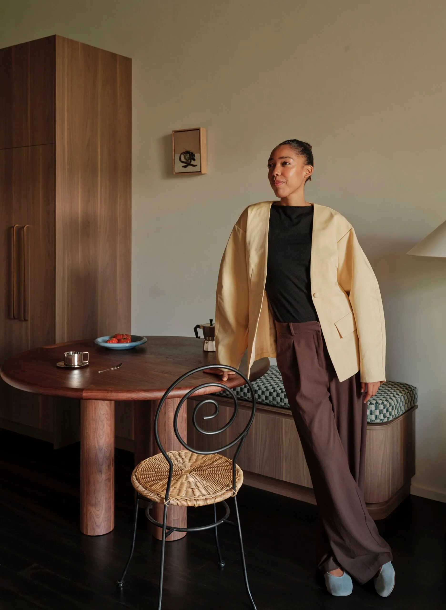 an Australian designer in a yellow jacket and brown pants leans on a table in a stylish room with wooden decor and a decorative chair.