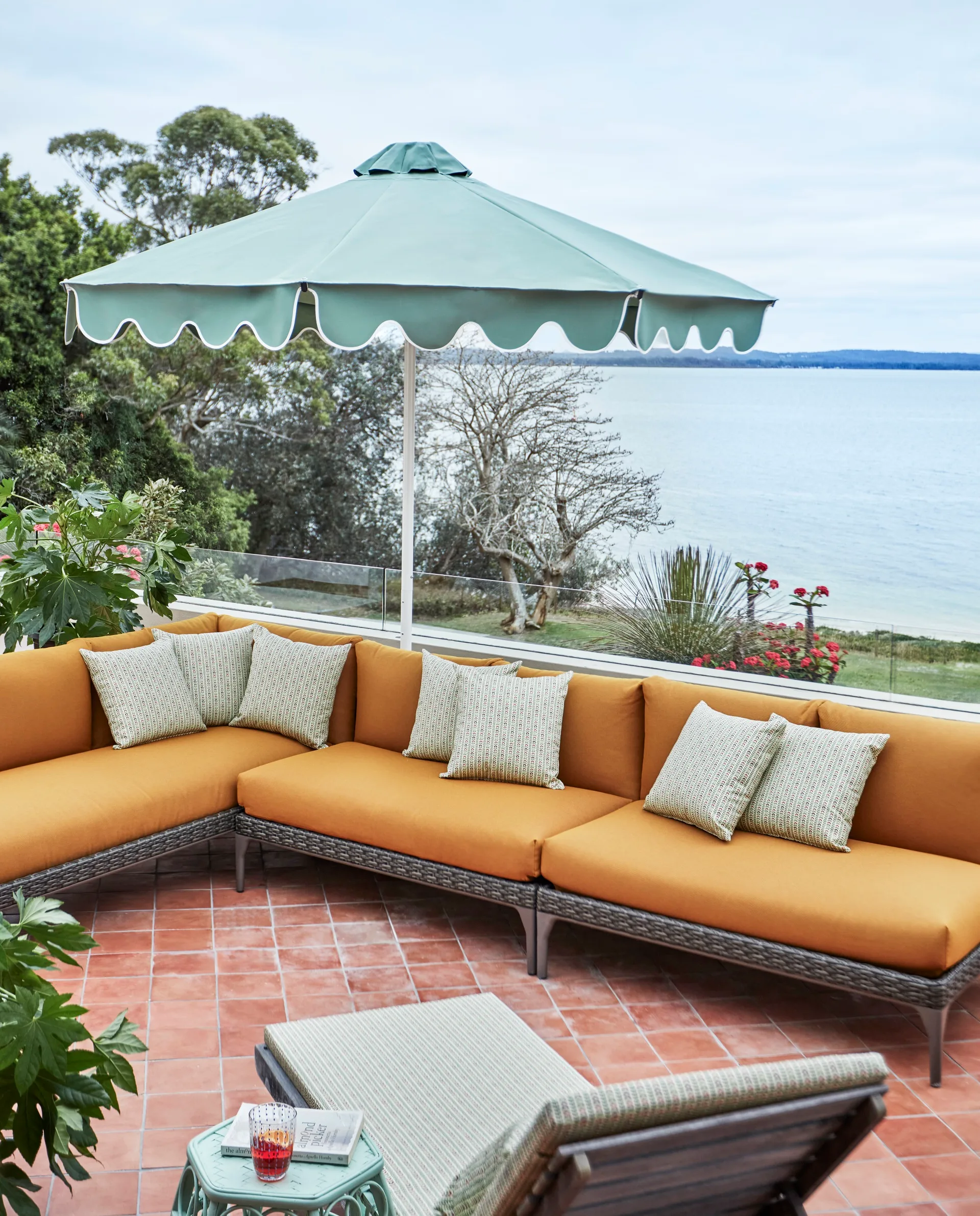 Outdoor patio with orange sectional sofa, green umbrella, and ocean view in the background. The terrace has a Mediterranean style with terracotta floor tiles.