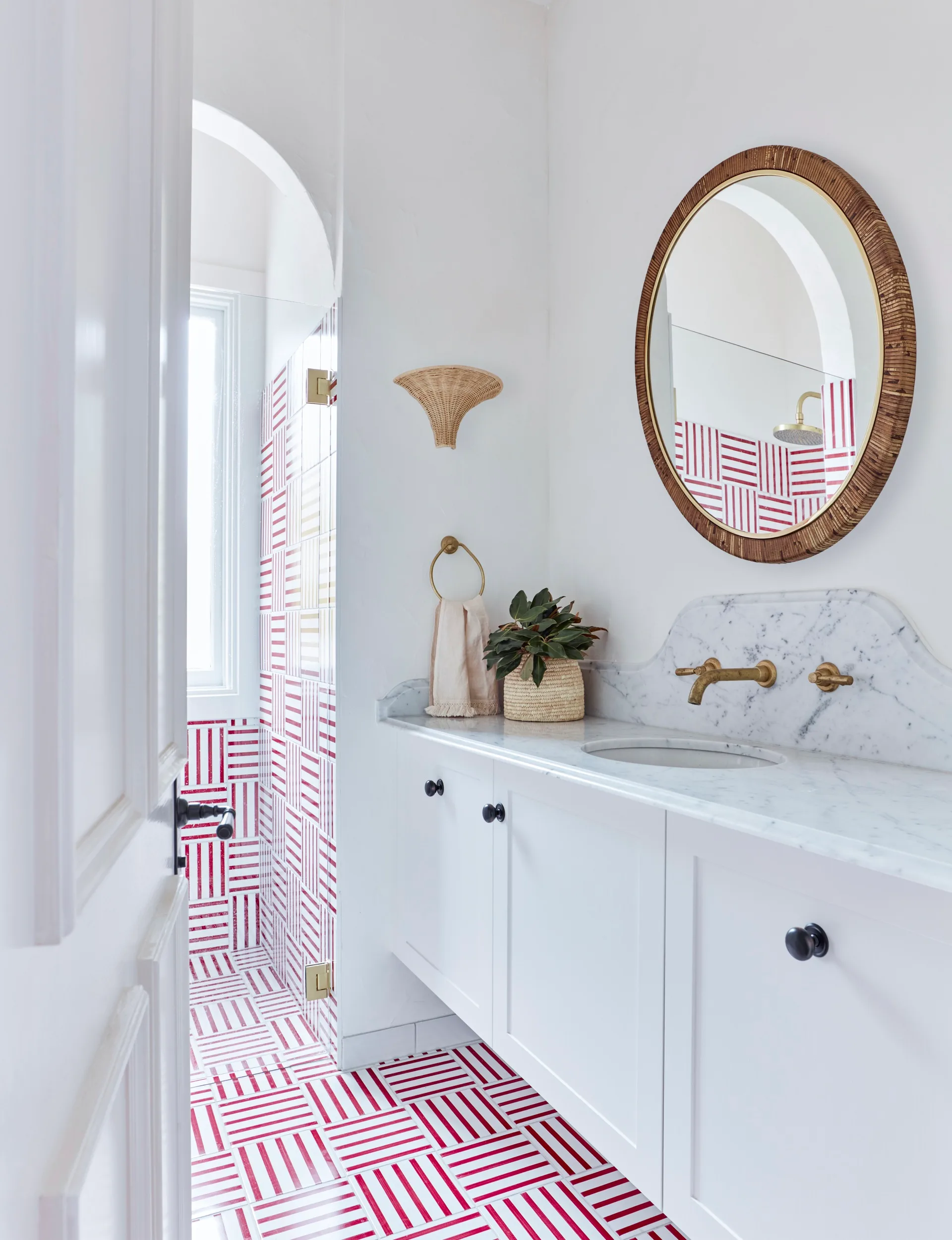 Red and white striped floor and wall tiles in a bathroom with round mirror, marble countertop, and brass fixtures. This vanity is in the foreground with the shower in the background.