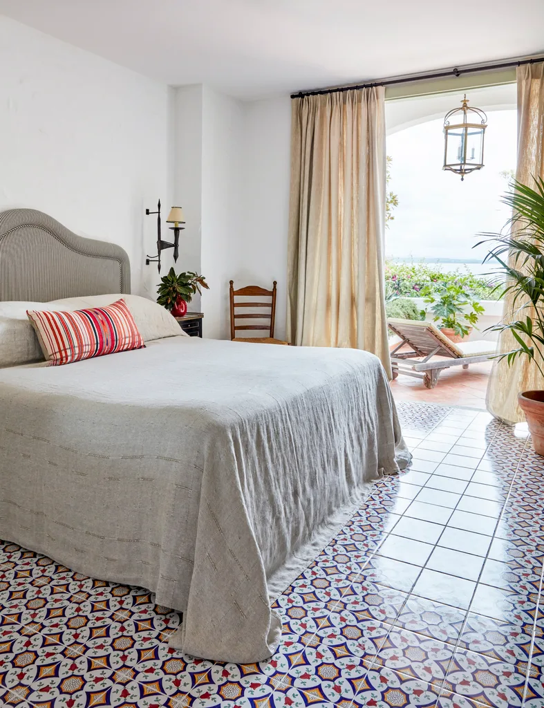 Cozy bedroom with patterned tiled floor, white bedspread, red-striped pillow, and door leading to a sunny terrace with plants and a sunlounger.