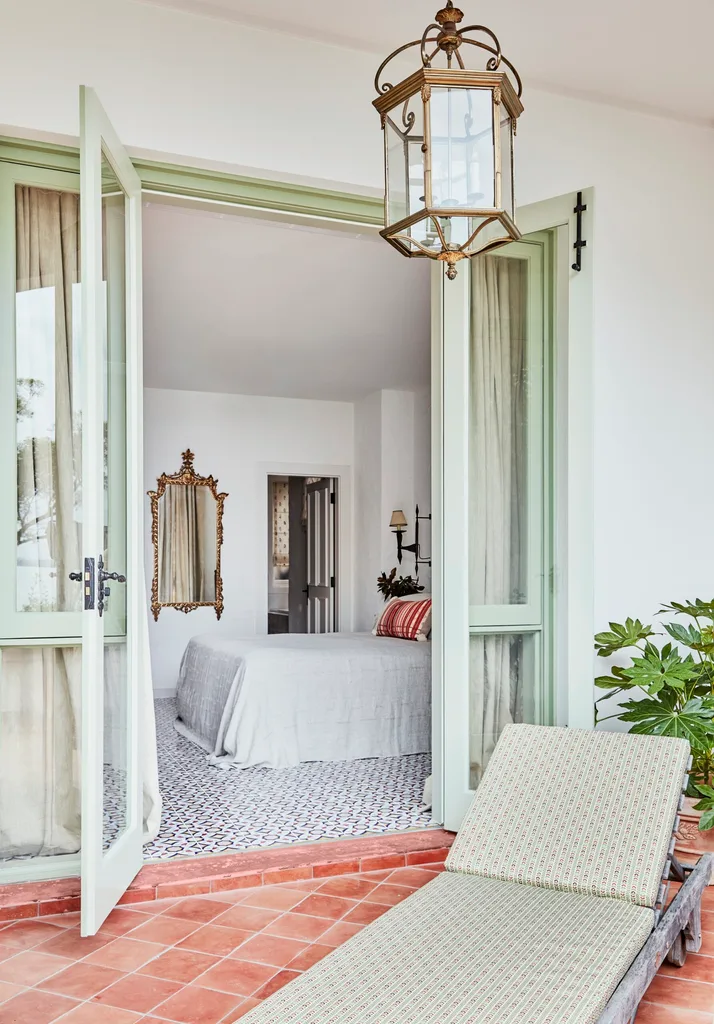 Stylish bedroom with open French doors leading to a terrace, vintage mirror, light gray bedding, and a patterned tile floor.