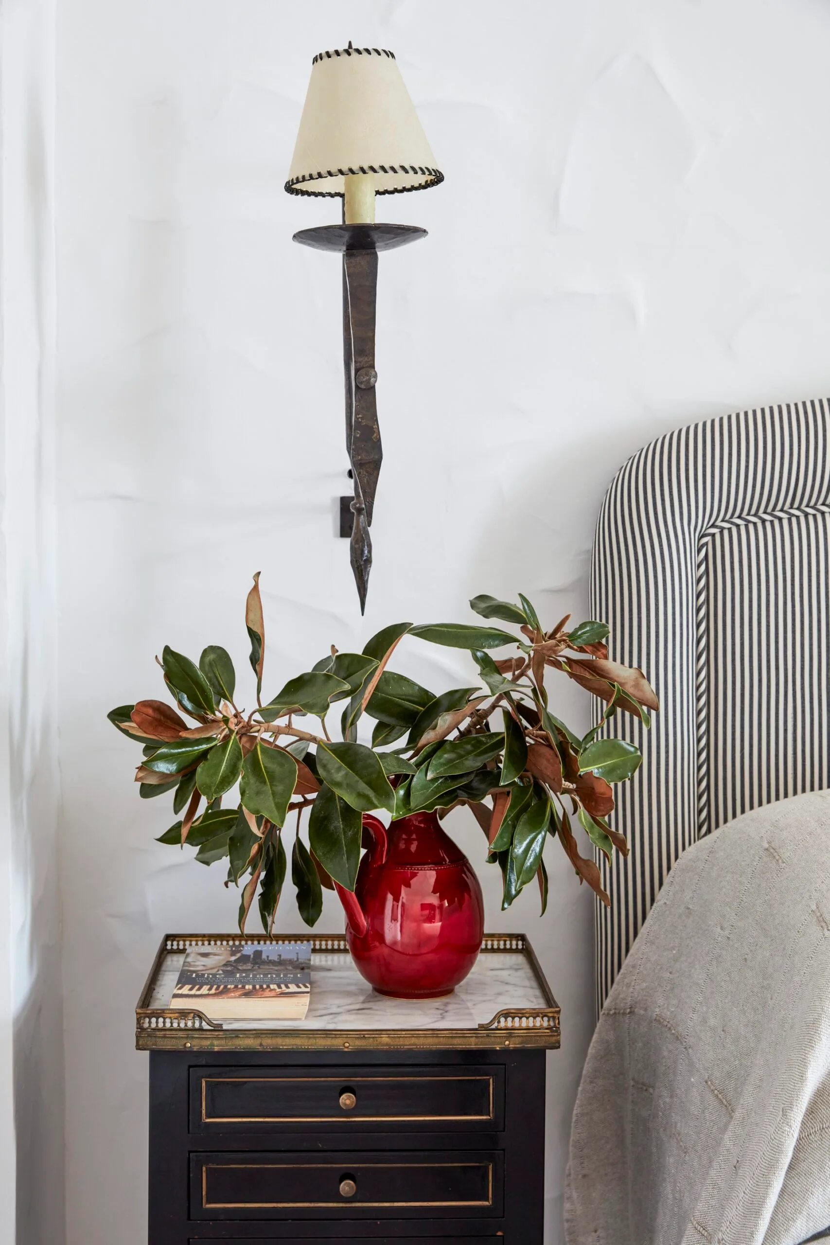 Nightstand with a red vase holding green leaves, a wall sconce with a lampshade, and a book in a bedroom setting.