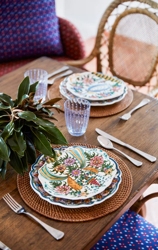 Elegantly set dining table with floral-patterned plates, blue glasses, and a leafy centerpiece on woven placemats.