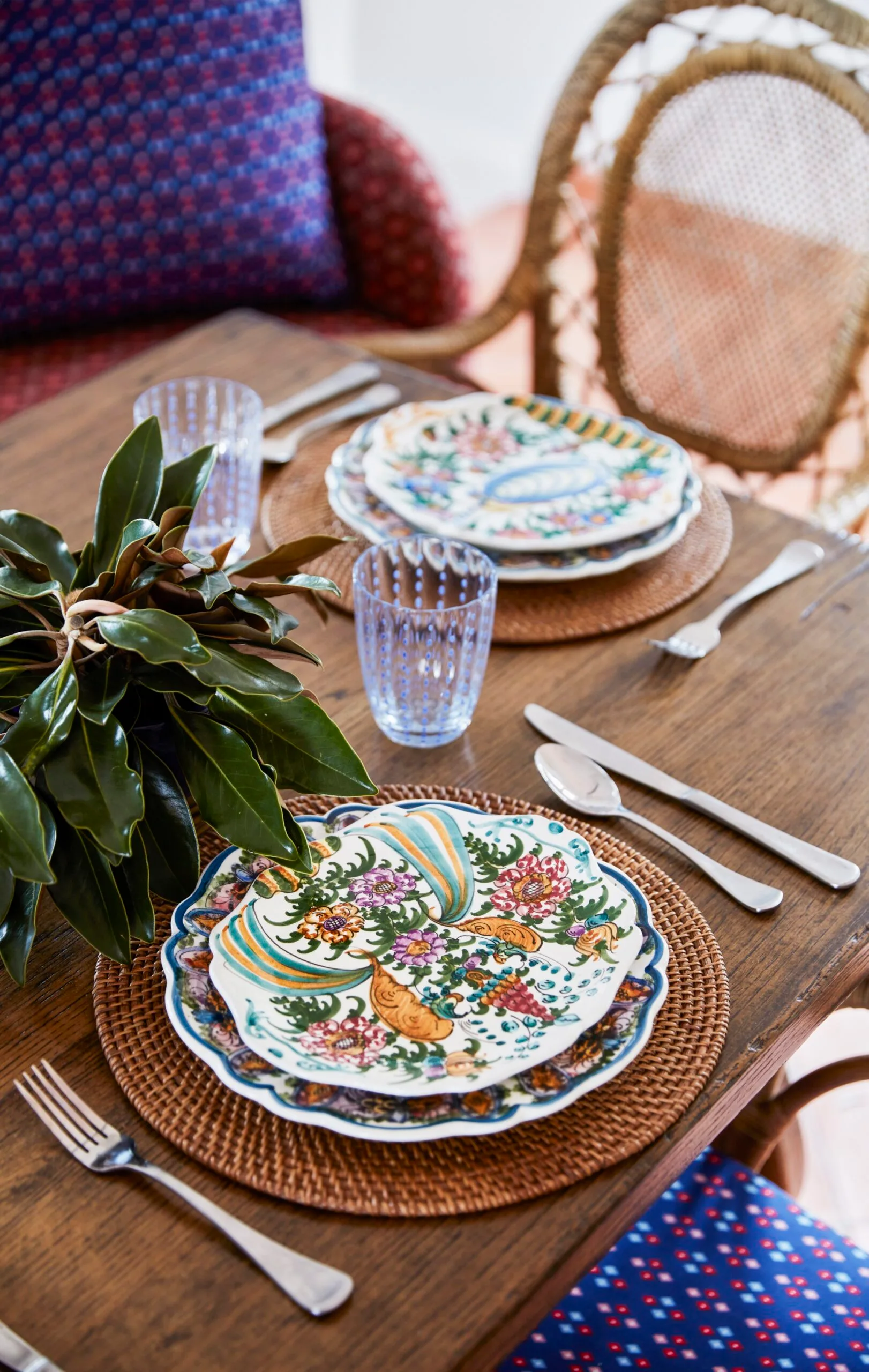 Elegantly set dining table with floral-patterned plates, blue glasses, and a leafy centerpiece on woven placemats.