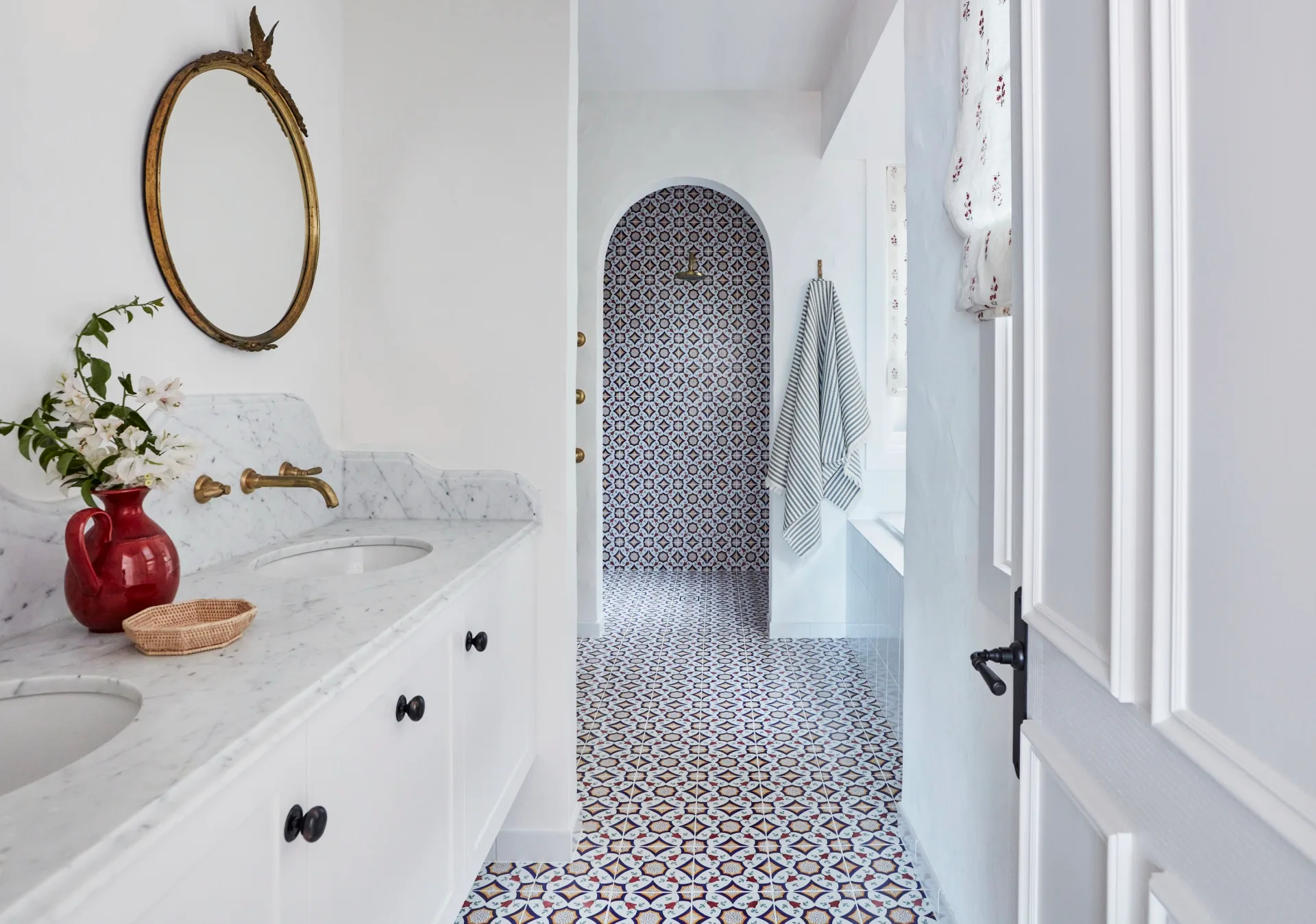 Elegant bathroom with marble counters, jack and jill basins, round mirror with gilded frame above the vanity, and patterned tile floor. The vanity is in the foreground and the background has a shower, visible through an arched open doorway. The floor tiles carry up the wall in the shower area.