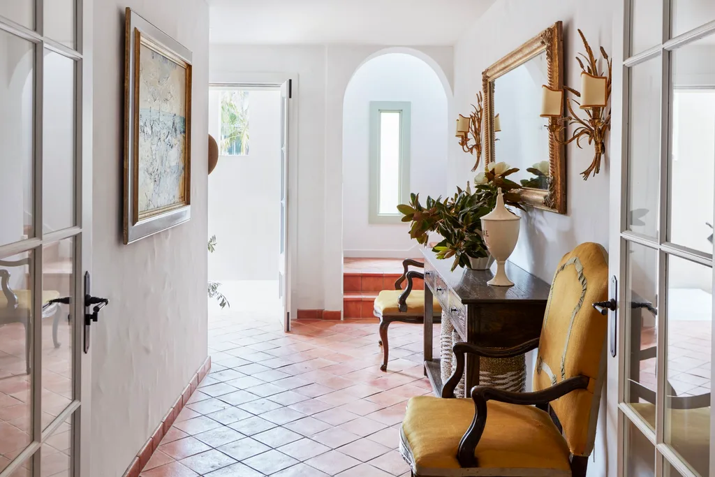 Hallway with terracotta tiles, antique yellow chairs, a mirror, wall sconces, and a painting. Bright natural light from open door. The style is Mediterranean.