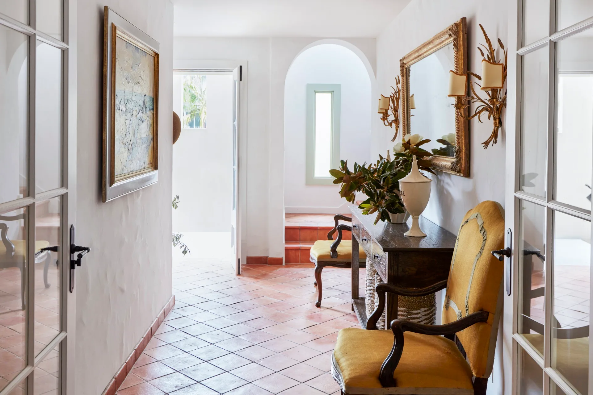Hallway with terracotta tiles, antique yellow chairs, a mirror, wall sconces, and a painting. Bright natural light from open door. The style is Mediterranean.