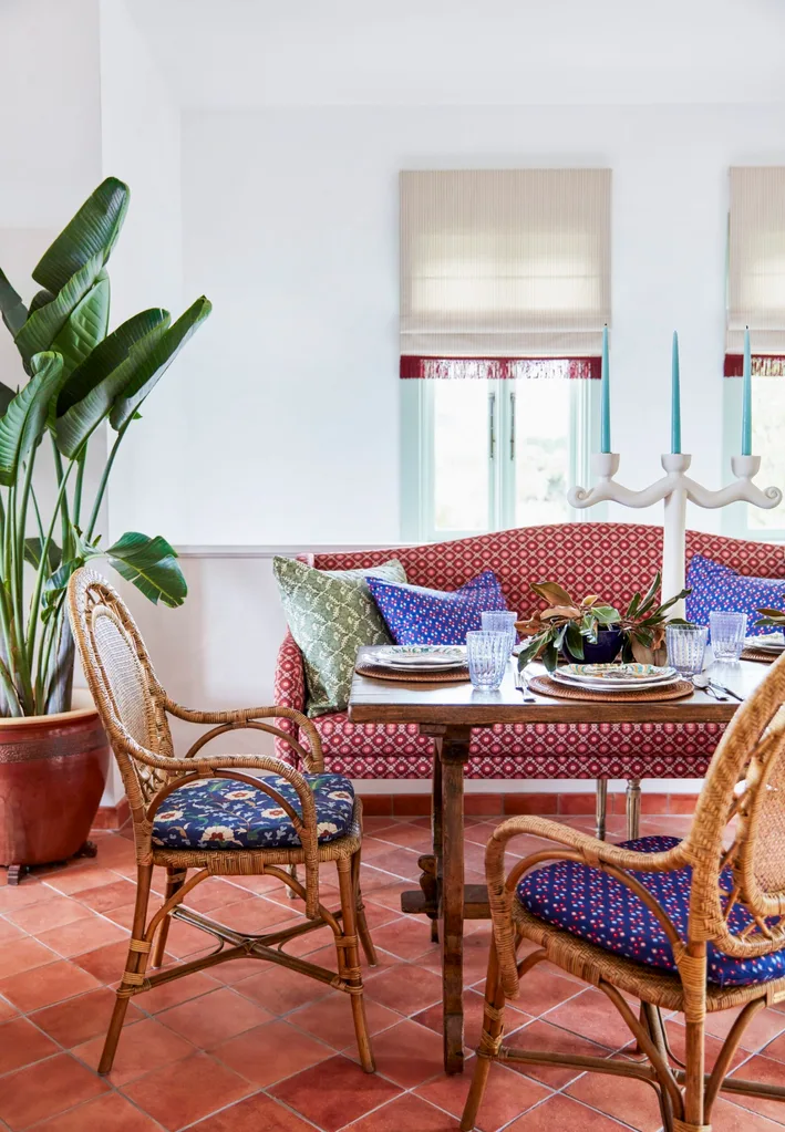 Bright dining area with a red patterned sofa that is a freestanding banquette, wicker chairs, a wooden table, potted plant, and blue candlesticks.