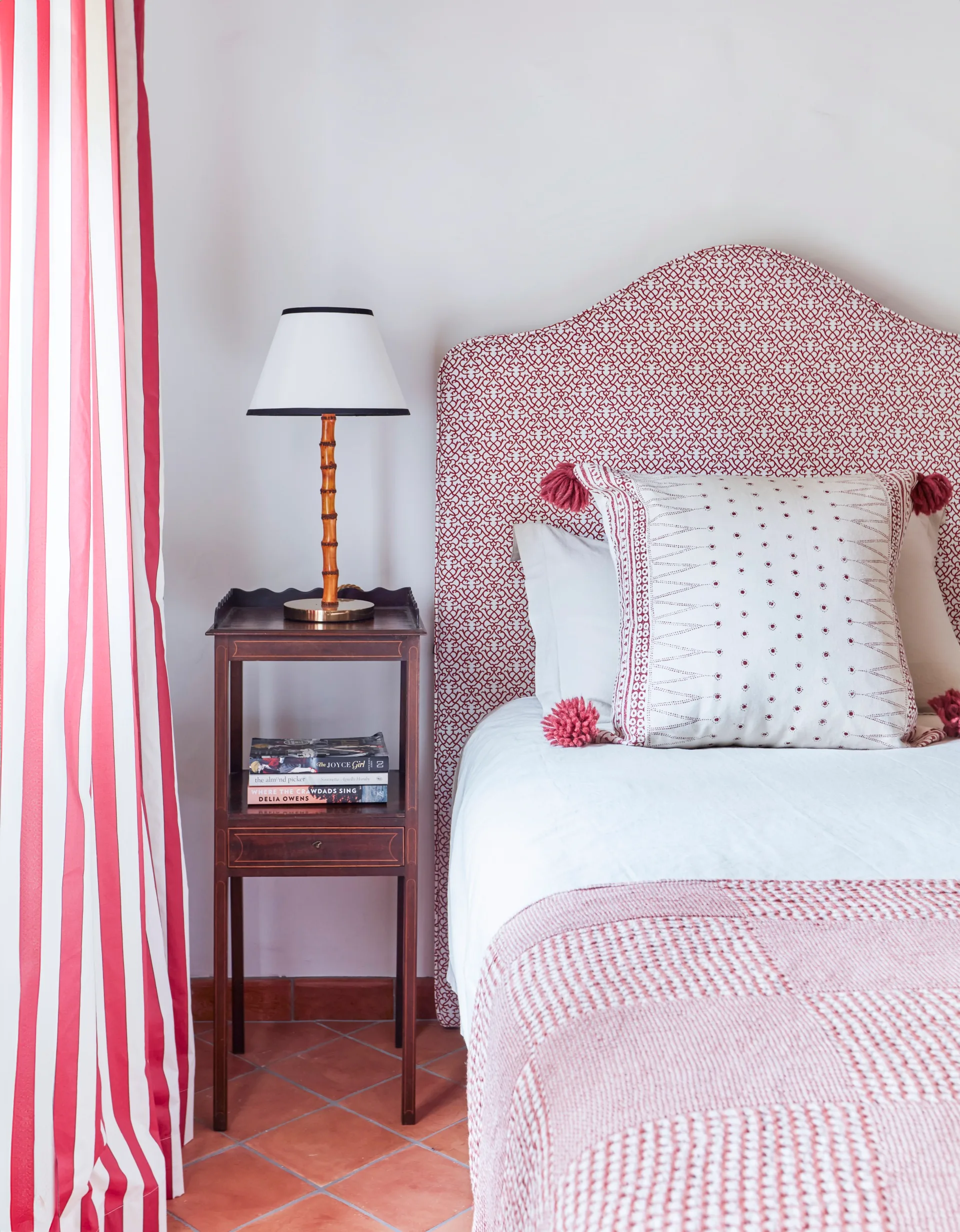 Bed with red patterned headboard, side table, lamp, and books; striped curtain on left.