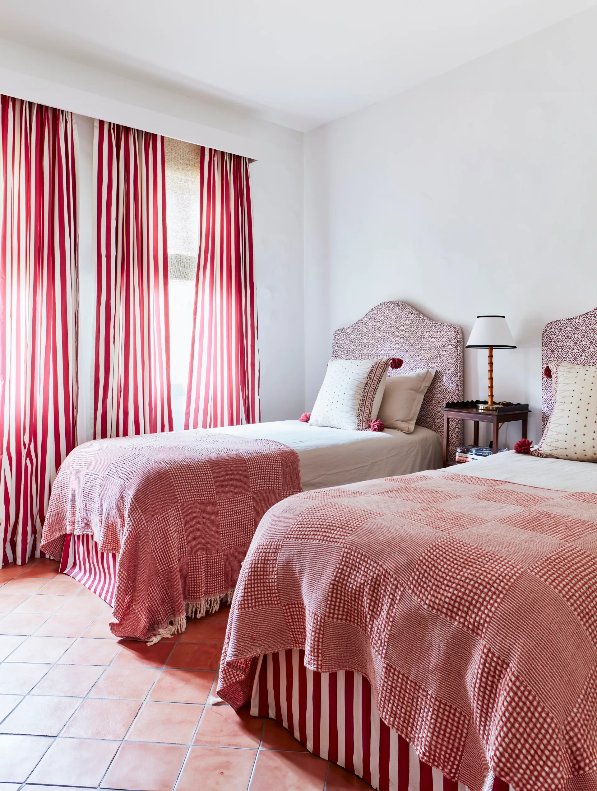 Two twin beds with red blankets, red and white striped curtains, and a small side table with a lamp in between the beds. The flooring is terracotta tiles.