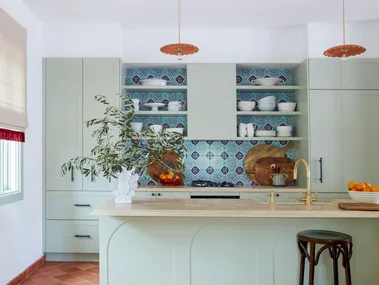 Modern kitchen with green cabinets, colorful tiled backsplash, pendant lights, and terracotta floor tiles.