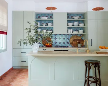 Modern kitchen with green cabinets, colorful tiled backsplash, pendant lights, and terracotta floor tiles.