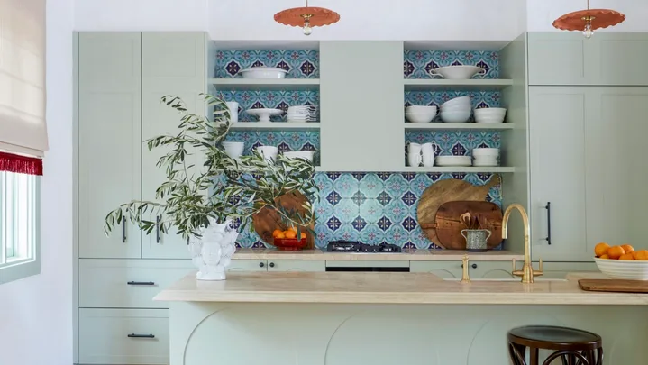 Modern kitchen with green cabinets, colorful tiled backsplash, pendant lights, and terracotta floor tiles.