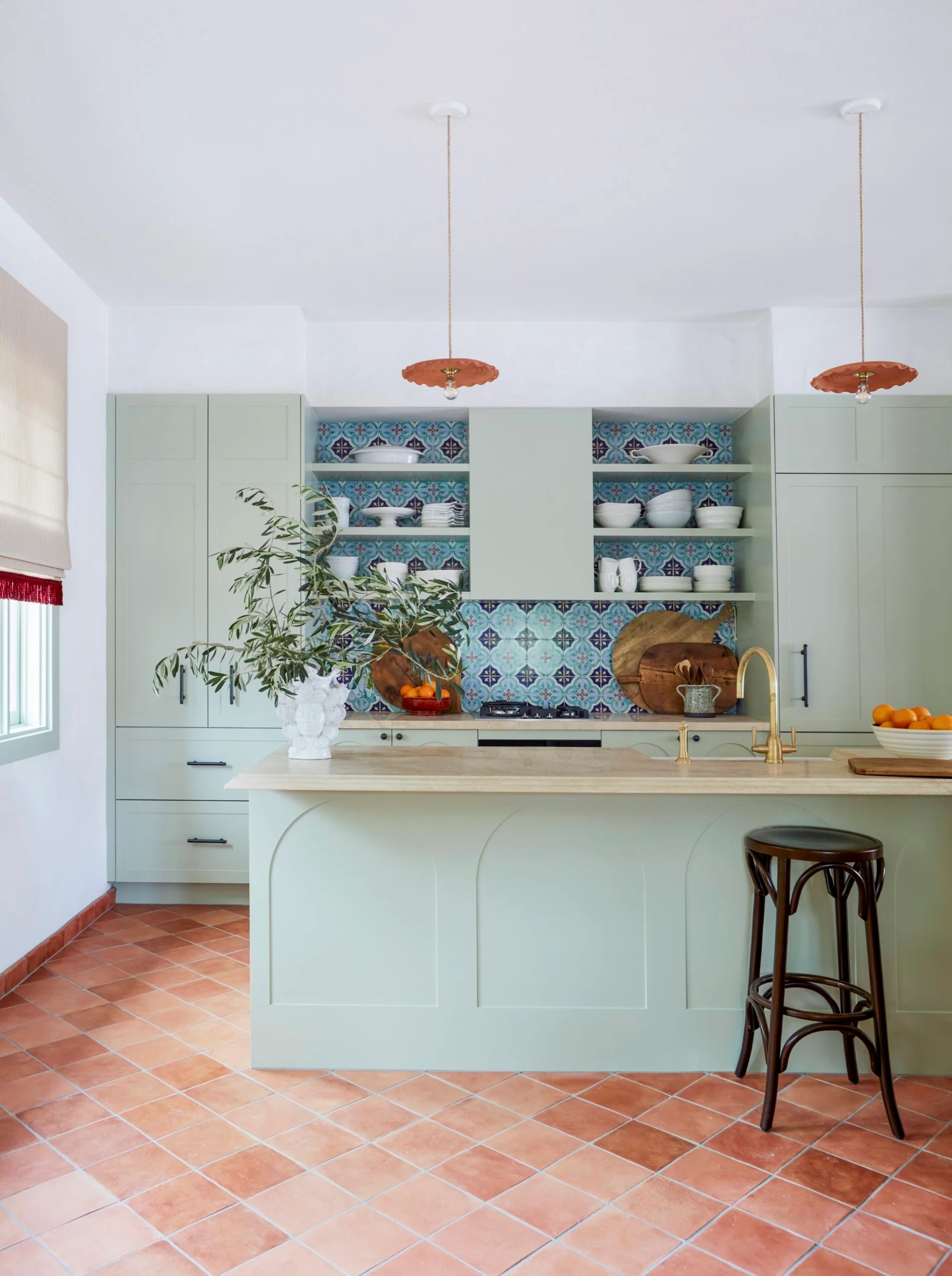 Modern kitchen with green cabinets, colorful tiled backsplash, pendant lights, and terracotta floor tiles.