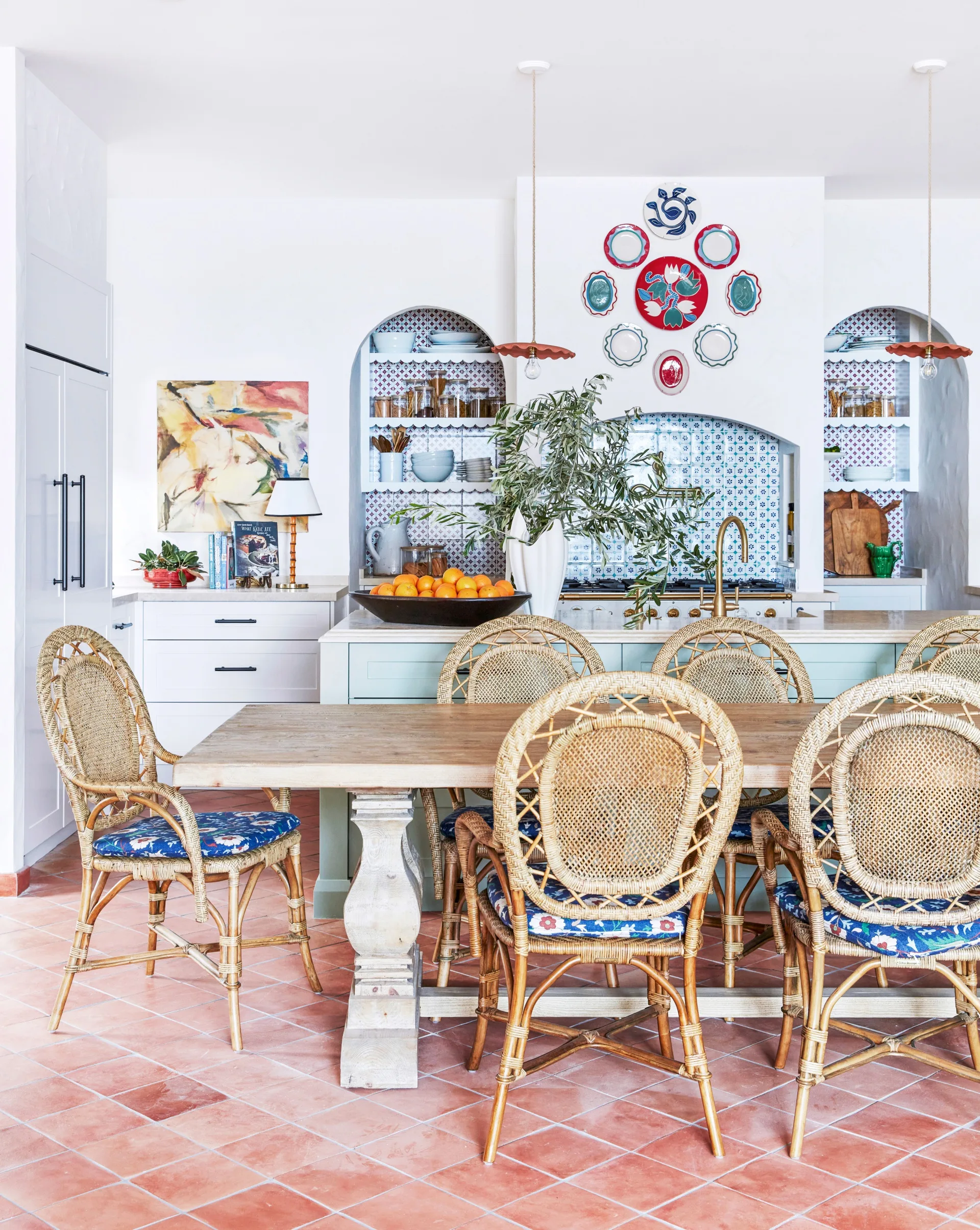 Cosy kitchen with wicker chairs, wooden dining table, open shelves, colorful decor, and a bowl of oranges on the island. The terracotta tiled floor adds character and a sense of Mediterranean style.