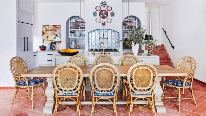 Decorative kitchen with rattan chairs, wooden table, terracotta tiled floor, and blue tile backsplash in arched alcoves, surrounded by white walls.