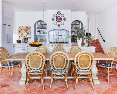 Decorative kitchen with rattan chairs, wooden table, terracotta tiled floor, and blue tile backsplash in arched alcoves, surrounded by white walls.
