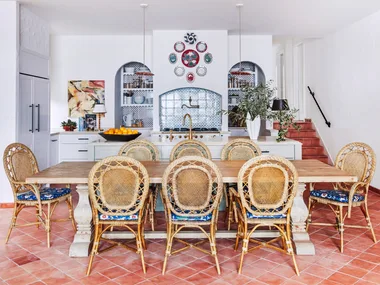 Decorative kitchen with rattan chairs, wooden table, terracotta tiled floor, and blue tile backsplash in arched alcoves, surrounded by white walls.