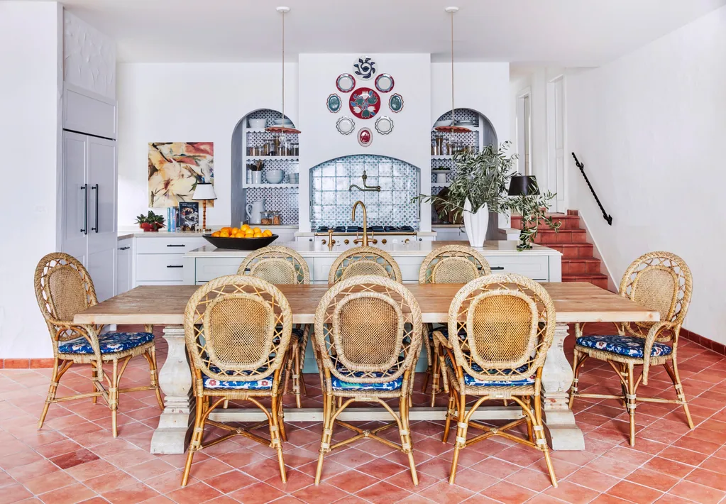 Decorative kitchen with rattan chairs, wooden table, terracotta tiled floor, and blue tile backsplash in arched alcoves, surrounded by white walls.