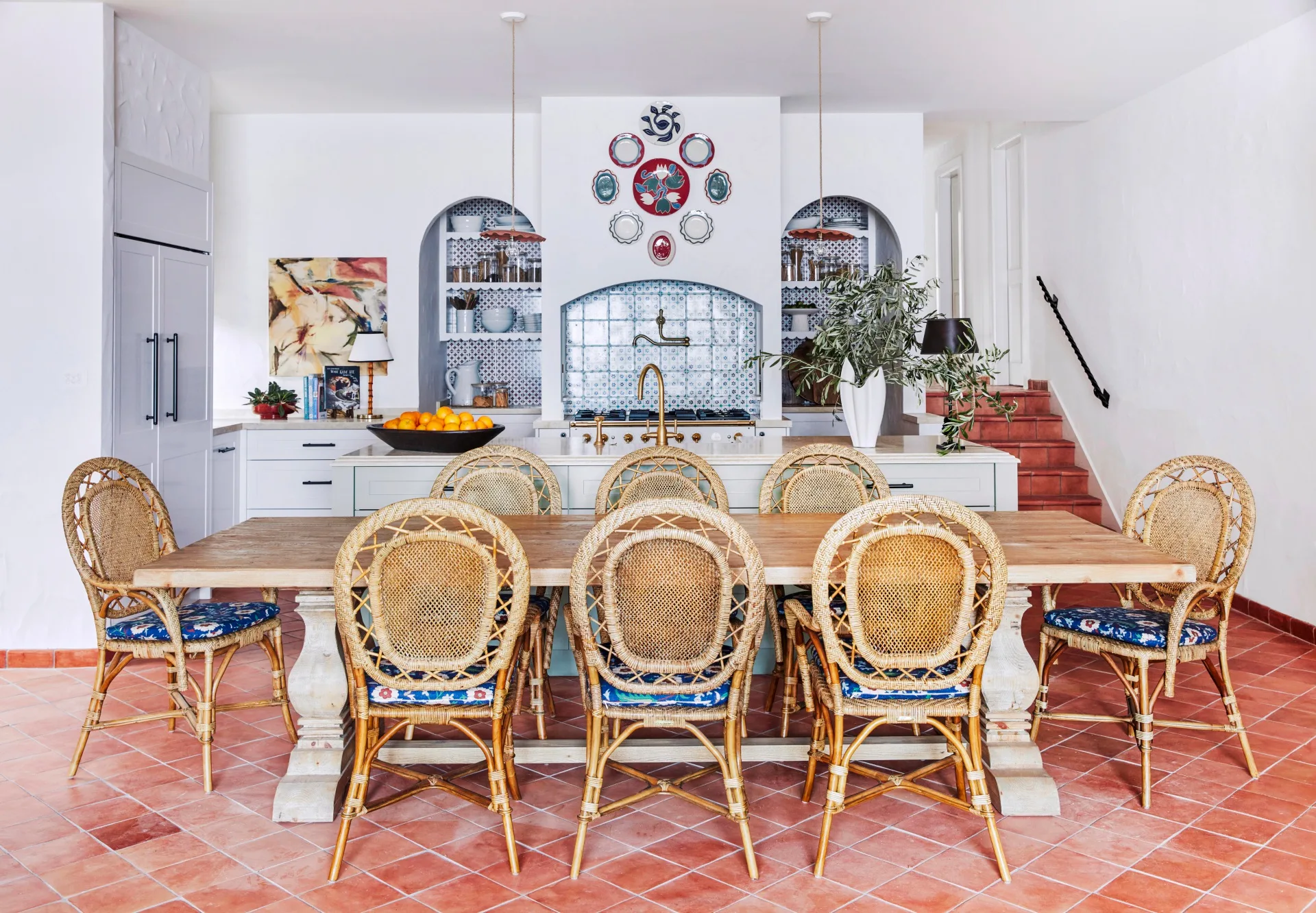 Decorative kitchen with rattan chairs, wooden table, terracotta tiled floor, and blue tile backsplash in arched alcoves, surrounded by white walls.