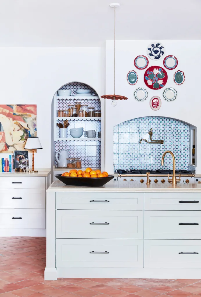 Chic kitchen with white cabinets, patterned splashback tiles in arched alcoves, oranges in a bowl on a sizable kitchen island, and decorative plates on the wall. This kitchen has a Mediterranean style.