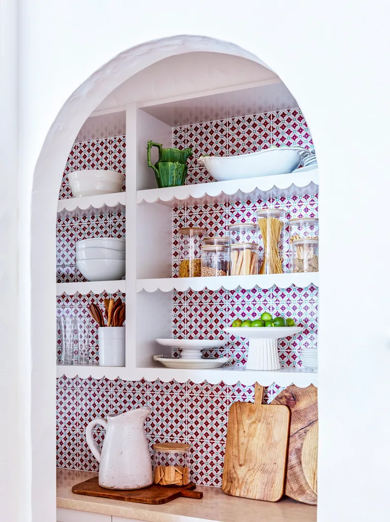 Open kitchen shelves with colorful tiled wall, holding dishes, jars, a green pitcher, wooden utensils, and cutting boards. The shelves have scalloped profiles.