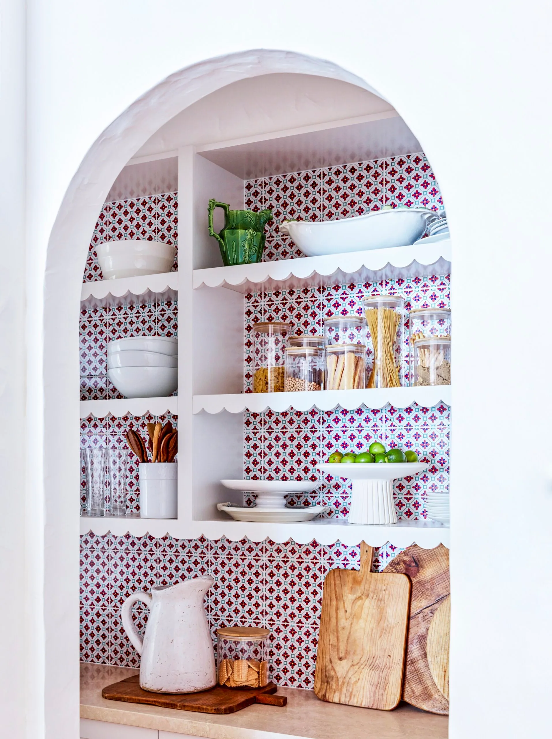 Open kitchen shelves with colorful tiled wall, holding dishes, jars, a green pitcher, wooden utensils, and cutting boards. The shelves have scalloped profiles.