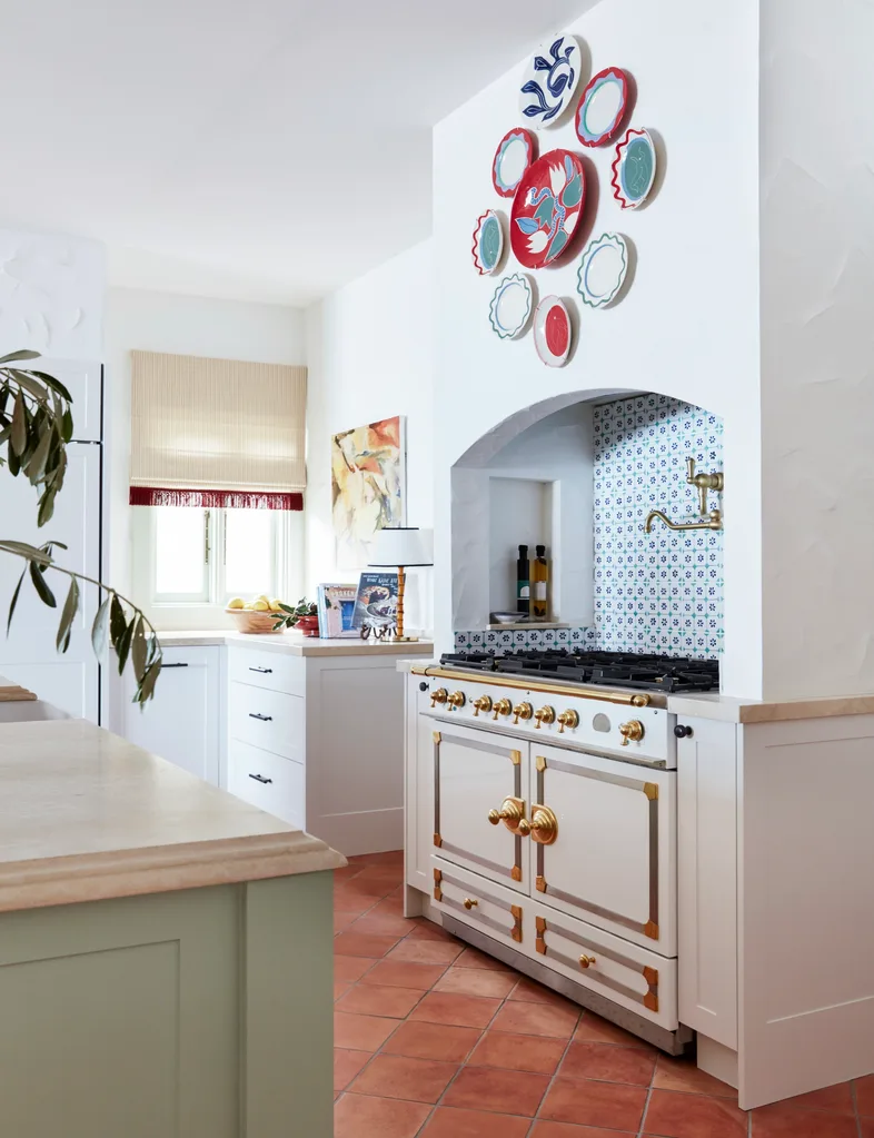 Large kitchen with white walls, white cabinetry, decorative plates on the wall, and terracotta tile flooring. A classic and large La Cornue oven and stovetop is positioned with a built-in rangehood above, in an arched alcove with a tiled splashback. Brass hardware includes a pot filler. The style is Mediterranean.