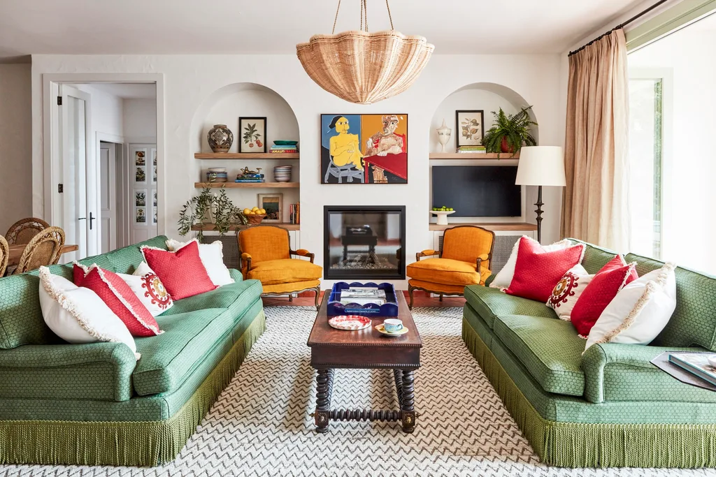 Cosy living room with green sofas, red and white cushions, and a central coffee table. Art and decor on open shelves inside arched alcoves flank a central fireplace and large artwork. A zig-zig patterned rug that is very large cover the terracotta tiled flooring. The space has a Mediterranean style vibe.